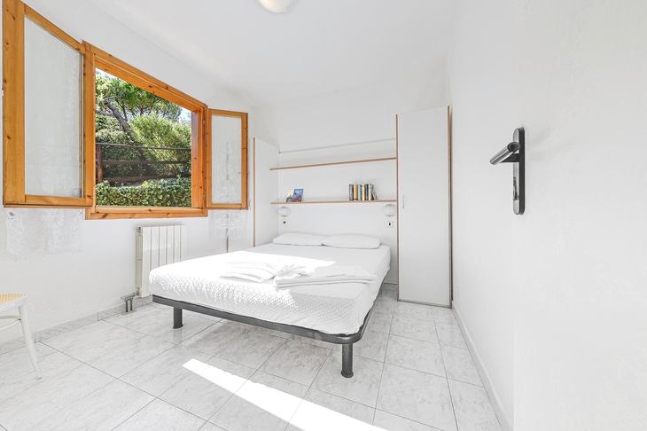 Helles Schlafzimmer mit Doppelbett, offenem Fenster, weißen Wänden und Fliesenboden.