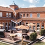Rotes rechtwinkliges Backsteingebäude der Ferienanlage Büsum mit Terrasse, Sitzmöbeln und Sonnenschirmen bei sonnigem Wetter.