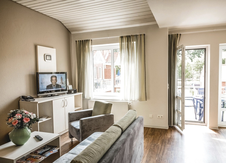 Wohnzimmer in Ferienwohnung mit Sofa, Sessel, Fernseher und großem Fenster zum Balkon.