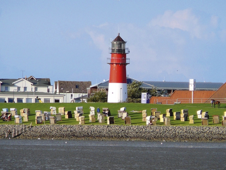 Leuchtturm auf einer Wiese am Meer von Büsum mit Strandkörben im Vordergrund, Häuser und blauer Himmel im Hintergrund.