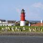 Leuchtturm auf einer Wiese am Meer von Büsum mit Strandkörben im Vordergrund, Häuser und blauer Himmel im Hintergrund.