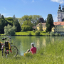 Radfahrer macht Pause am Flussufer mit Blick auf eine Kirche und Gebäude im Hintergrund.