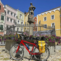 Rotes Fahrrad vor einem Brunnen auf dem Tauernradweg in einer Stadtumgebung.