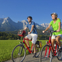 Zwei Radfahrer auf einem Weg vor Bergpanorama bei sonnigem Wetter.
