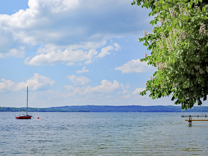 Blick auf den Starnberger See mit Boot, Bäumen und bewölktem Himmel.