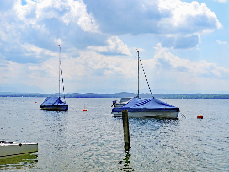 Boote schwimmen auf dem Starnberger See vor einer Alpenkulisse unter bewölktem Himmel.