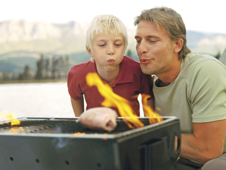 Ein Mann und ein Kind pusten auf einem Grill ins Feuer.