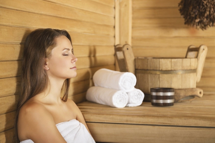 Frau in Sauna mit Holzverkleidung, weißen Handtüchern und Wasserbottich.