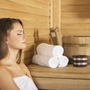 Frau in Sauna mit Holzverkleidung, weißen Handtüchern und Wasserbottich.