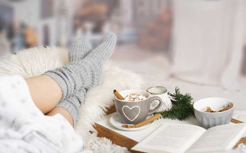 Füße in grauen Socken auf Fell, Tee und Buch im Hintergrund, winterlich gemütlich.