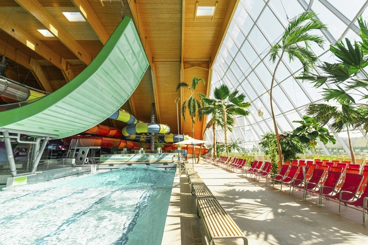 Innenansicht einer Therme mit Rutschen, Palmen und Liegestühlen am Beckenrand.