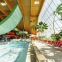 Innenansicht einer Therme mit Rutschen, Palmen und Liegestühlen am Beckenrand.