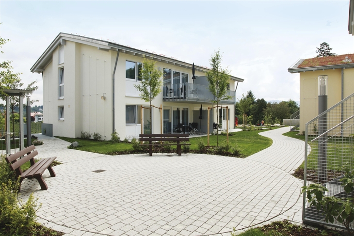 Modernes zweistöckiges Gebäude mit gepflastertem Weg und Bänken in einer Grünanlage.