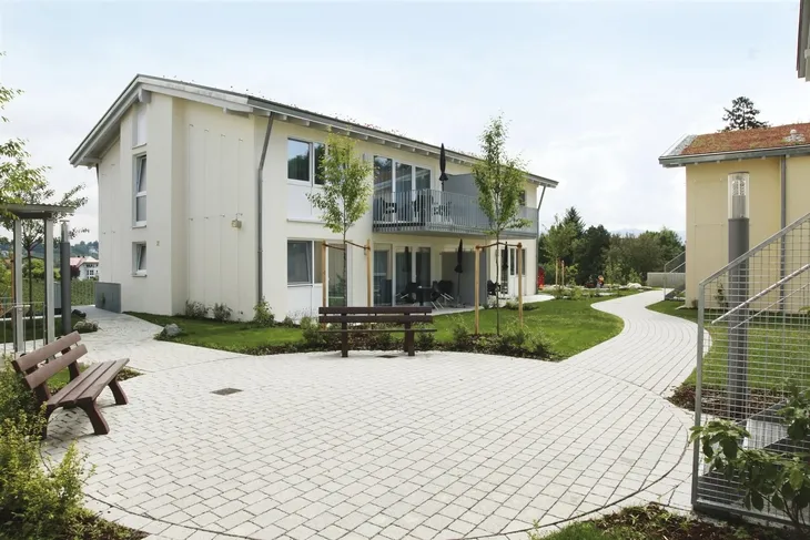 Modernes zweistöckiges Gebäude mit gepflastertem Weg und Bänken in einer Grünanlage.
