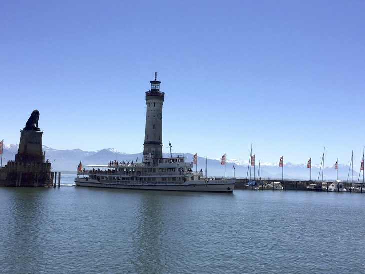 Ein Dampfschiff fährt vor einem Leuchtturm im Hafen, Berge im Hintergrund.
