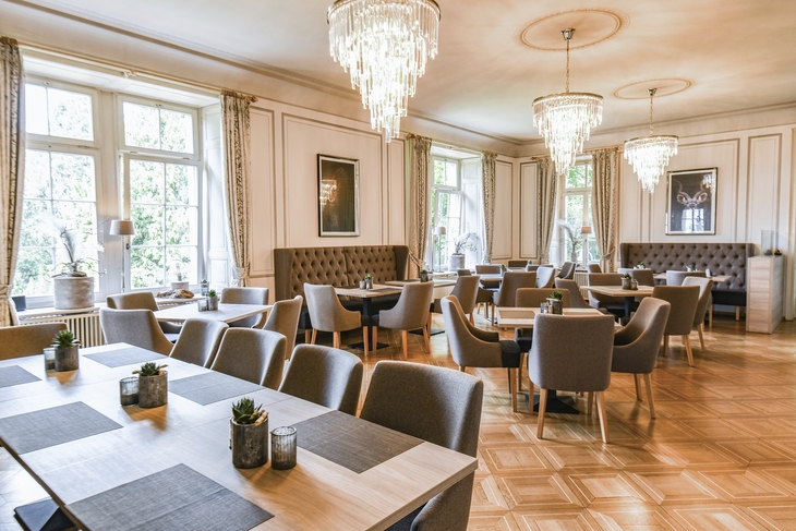 Elegantes Restaurantinterieur in der Villa Alwind in der Ferienanlage Lindau. Unterschiedlich große Holztische mit grauen, gepolsterten Stühlen auf Holzboden, sowie große weiße Fenster mit zusammengebundenen Vorhängen und moderne Kronleuchter an der Decke, die das Restaurant erhellen.