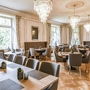 Elegantes Restaurantinterieur in der Villa Alwind in der Ferienanlage Lindau. Unterschiedlich große Holztische mit grauen, gepolsterten Stühlen auf Holzboden, sowie große weiße Fenster mit zusammengebundenen Vorhängen und moderne Kronleuchter an der Decke, die das Restaurant erhellen.