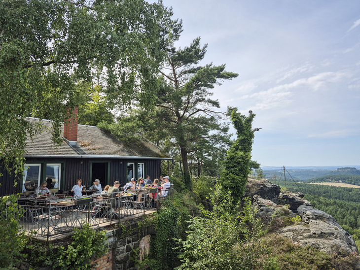 Terrasse einer Bergwirtschaft mit Gästen, Blick auf bewaldete Landschaft und Fernsicht.