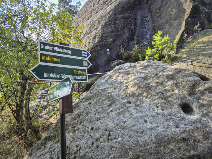 Wegweiser am Malerweg mit grünen Schildern und Felsen im Hintergrund.