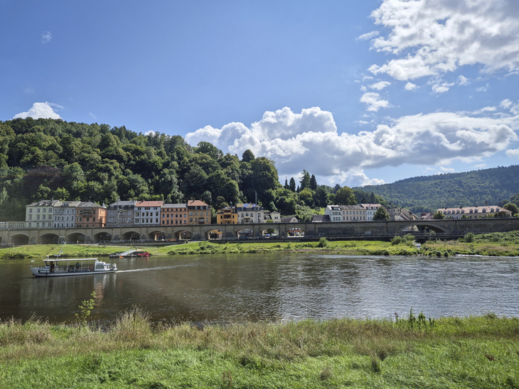 Flusslandschaft mit Boot, Wiese im Vordergrund, bunte Häuser und bewaldete Hügel im Hintergrund.