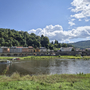 Flusslandschaft mit Boot, Wiese im Vordergrund, bunte Häuser und bewaldete Hügel im Hintergrund.