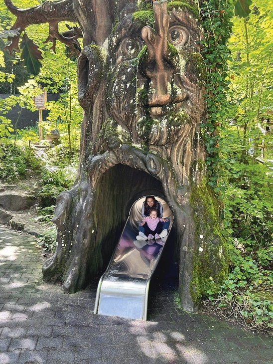 Person rutscht auf einer Rutsche, die aus einem Baumstamm mit Gesicht herausragt.
