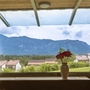 Blick aus einem Fenster auf Berge und Häuser, Vase mit Blumen im Vordergrund.