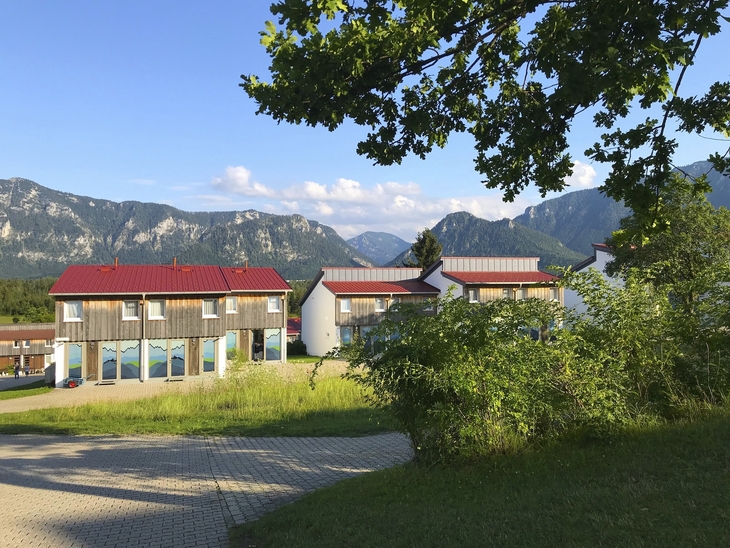 Gebäude vor Berglandschaft mit klarem Himmel und Bäumen im Vordergrund.