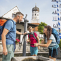 Drei Wanderer an einem Brunnen in Bayern vor einer Kirche und Maibaum.