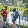Zwei Wanderer mit Rucksäcken schauen auf einen Fluss in einer bewaldeten Berglandschaft.