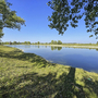 Blick auf einen See mit grasbewachsenem Ufer und Bäumen bei klarem Himmel.