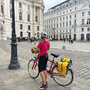 Frau mit Helm steht mit Fahrrad auf einem gepflasterten Platz vor historischen Gebäuden.
