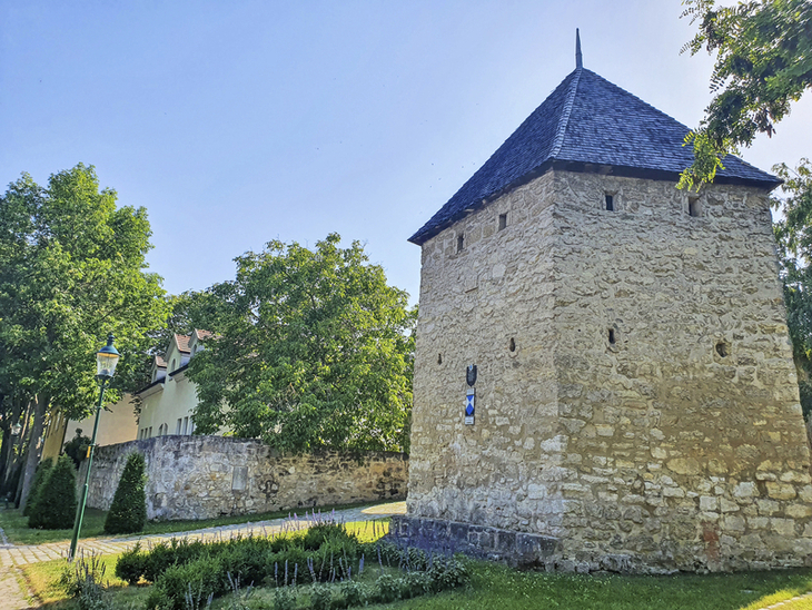 Steinerner Turm mit Spitzdach und grünen Bäumen im Hintergrund bei sonnigem Wetter.