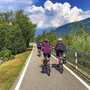 Radfahrer auf einem Radweg, umgeben von Bäumen und Bergen bei sonnigem Wetter.