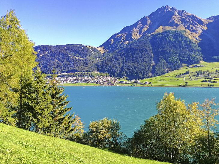 Reschensee mit Dorf am Ufer, umgeben von Bergen und grüner Landschaft.