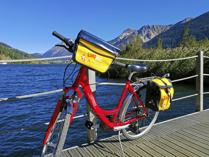 Rotes Fahrrad auf einem Steg am Haidersee mit Bergkulisse im Hintergrund.