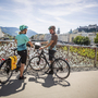 Zwei Radfahrer auf einer Brücke mit Blick auf eine Stadt im Hintergrund.