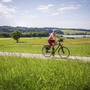 Radfahrerin auf einem Weg am Chiemsee mit Blick auf Wiesen und Wälder.