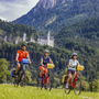 Drei Radfahrer vor Schloss Neuschwanstein in einer bergigen Landschaft.