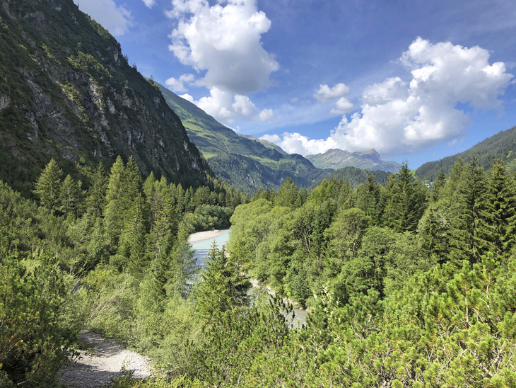 Berglandschaft mit Fluss und bewaldeten Hügeln unter blauem Himmel.