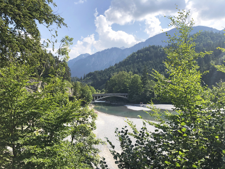 Blick auf einen Fluss in einem bewaldeten Tal mit Bergen im Hintergrund und einer Brücke.