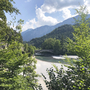 Blick auf einen Fluss in einem bewaldeten Tal mit Bergen im Hintergrund und einer Brücke.