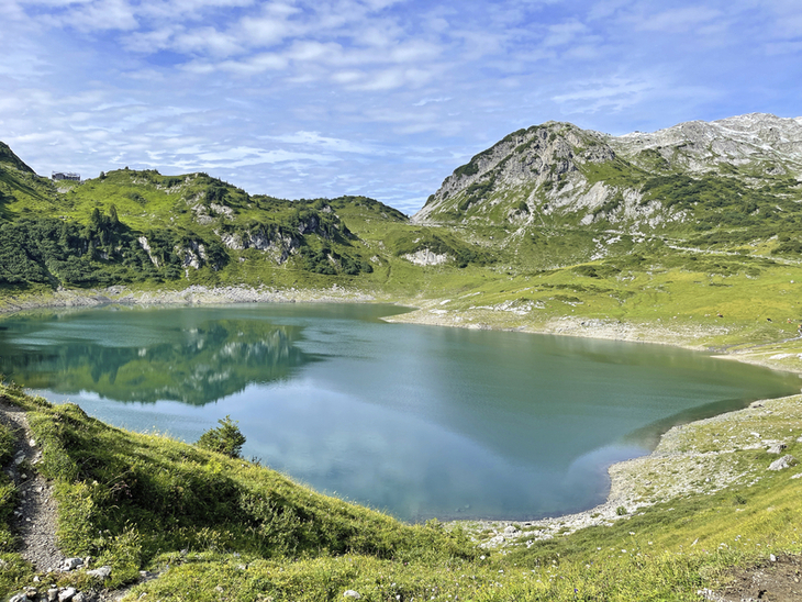 Bergsee mit grünem Ufer und bewachsenen Hügeln im Hintergrund unter blauem Himmel.