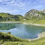 Bergsee mit grünem Ufer und bewachsenen Hügeln im Hintergrund unter blauem Himmel.