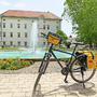 Fahrrad vor historischem Gebäude und Brunnen mit Blumenbeet im Vordergrund.