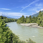 Fluss von Bäumen gesäumt, Berge im Hintergrund, blauer Himmel mit Wolken.