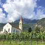 Kirche neben Weinreben mit Bergen im Hintergrund bei sonnigem Wetter.