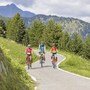 Drei Radfahrer auf einem Bergweg, umgeben von grünen Bäumen und Bergen.
