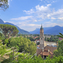 Blick auf Meran mit Kirche und umliegenden Bergen vom Tappeinerweg bei sonnigem Wetter.