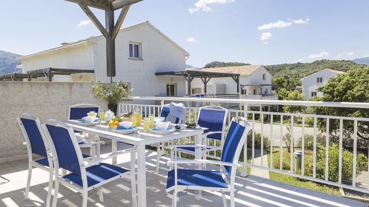 Terrasse mit Esstisch und blauen Stühlen, Blick auf Wohnhäuser und Hügel im Hintergrund.